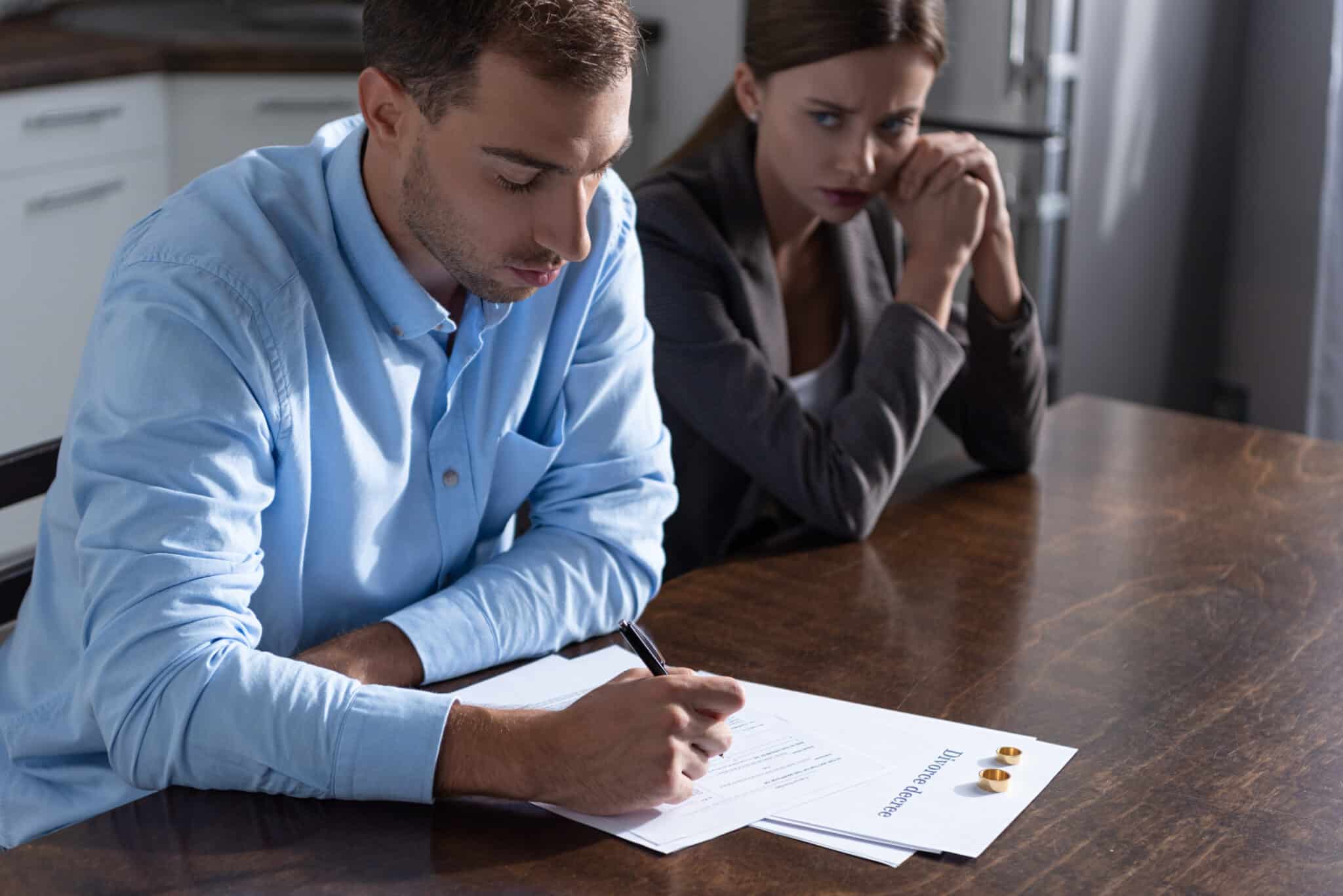  A man and a woman are sitting at a table. The man is signing divorce papers, and the woman is looking at him with a sad expression. There are wedding rings on the table.